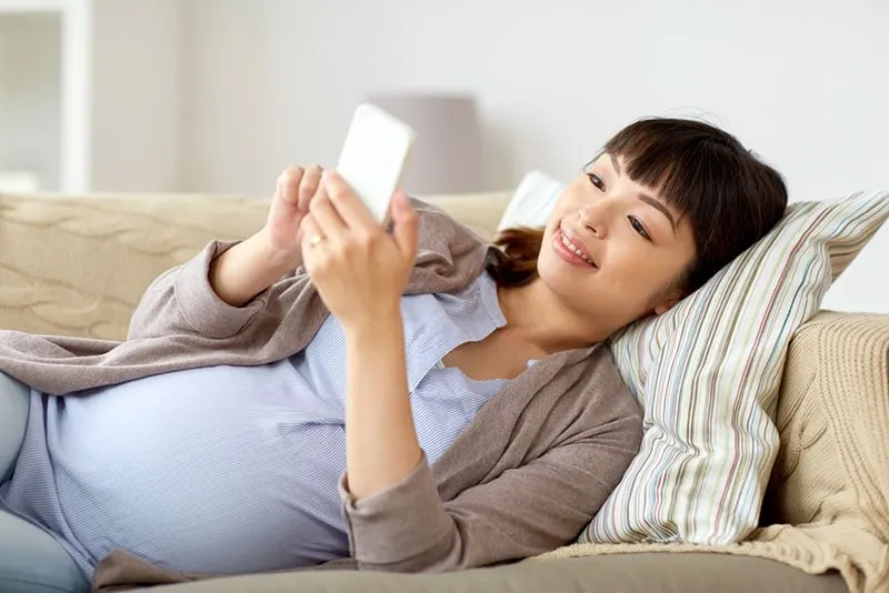 glückliche schwangere Frau, die auf der Couch liegt und Telefon ansieht