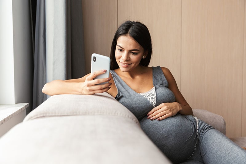 schöne schwangere Frau, die Telefon auf der Couch betrachtet
