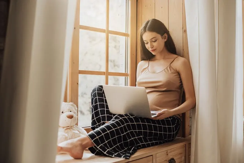 schöne schwangere Frau, die auf Laptop durch das Fenster tippt