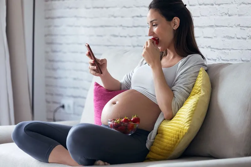 schwangere Frau, die Erdbeeren isst und Telefon ansieht