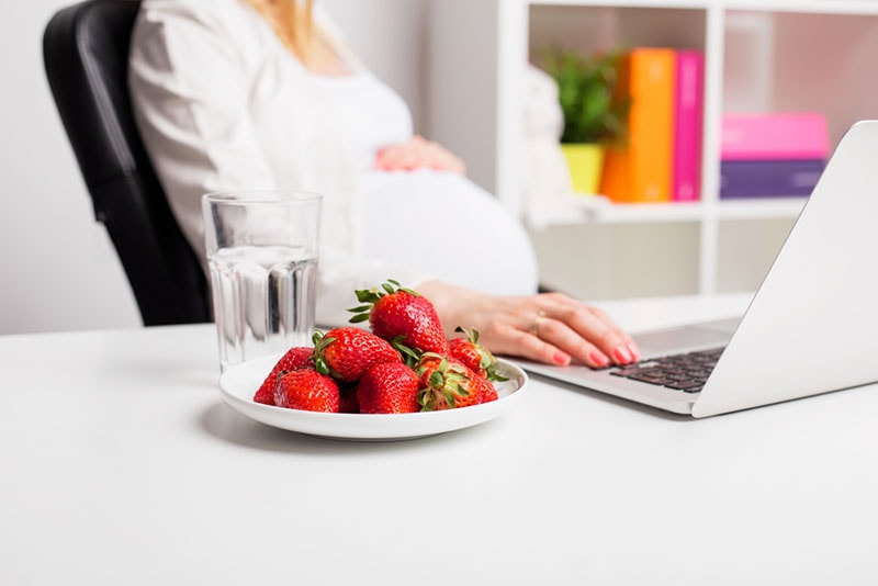 schwangere Frau, die auf Laptop rollt und Erdbeeren isst