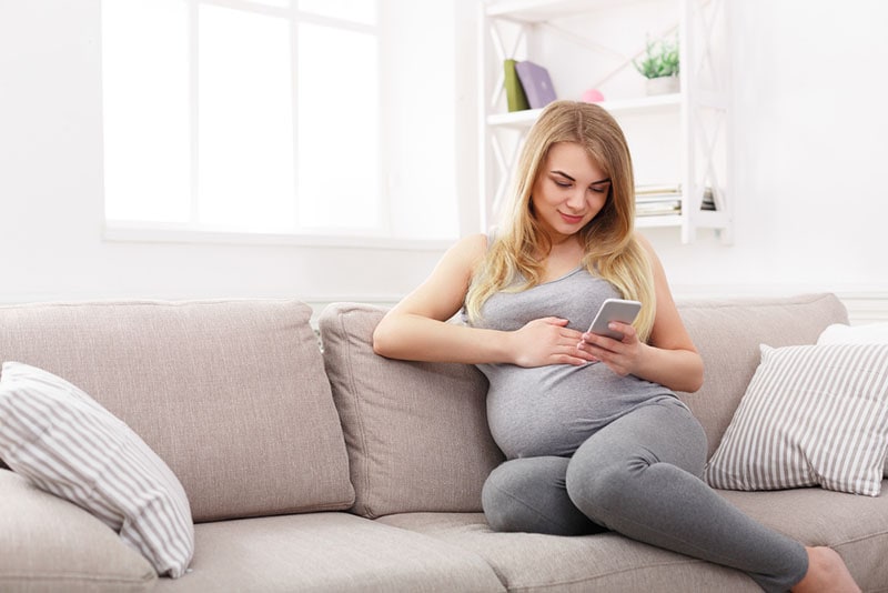 schwangere Frau, die auf der Couch sitzt und Telefon ansieht