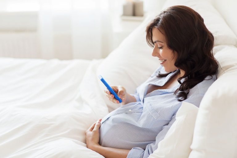 schwangere Frau, die Telefon im Bett betrachtet