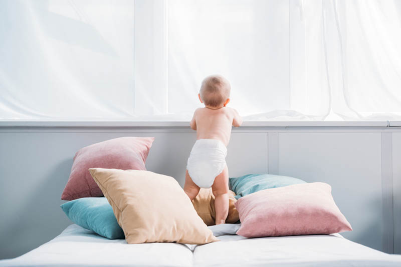 Baby trägt Windeln auf dem Bett, umgeben von bunten Kissen