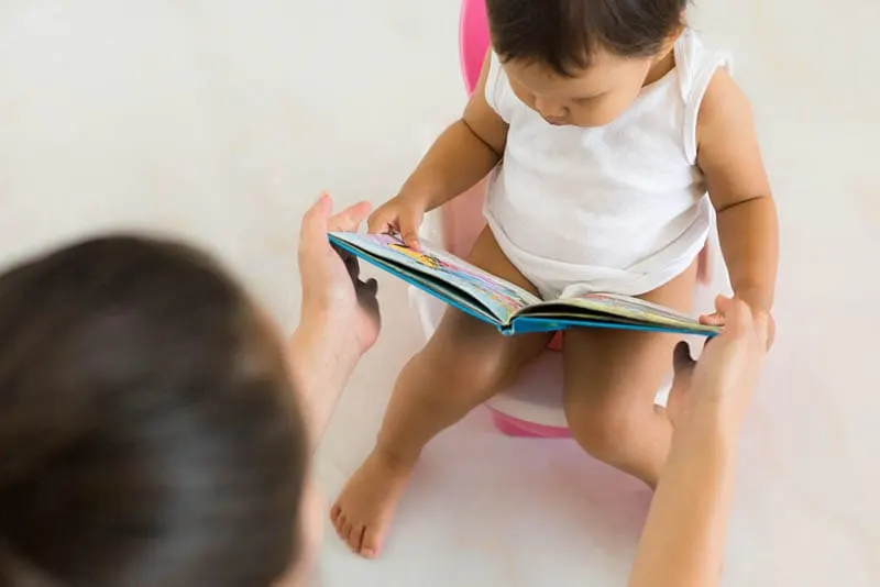 Mutter hält Buch an ein kleines Mädchen, während sie auf dem Töpfchen ist