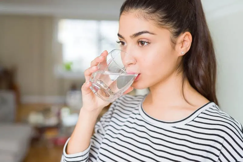 Schöne junge Frau, die ein frisches Glas Wasser zu Hause trinkt