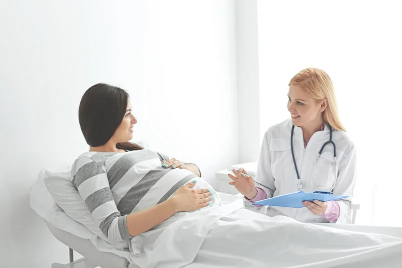 junge schwangere Frau, die auf Krankenhausbett liegt und mit einem Arzt spricht