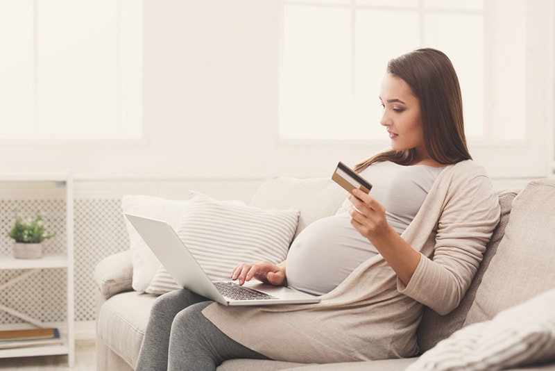 junge schwangere Frau, die auf der Couch im Wohnzimmer mit Kreditkarte in der Hand sitzt