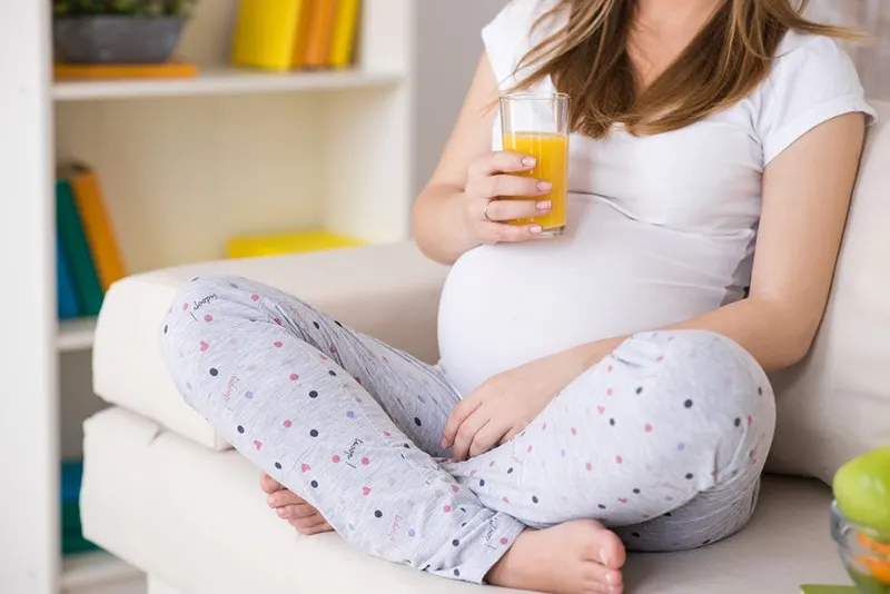 junge schwangere Frau, die auf der Couch sitzt und orange Flüssigkeit trinkt