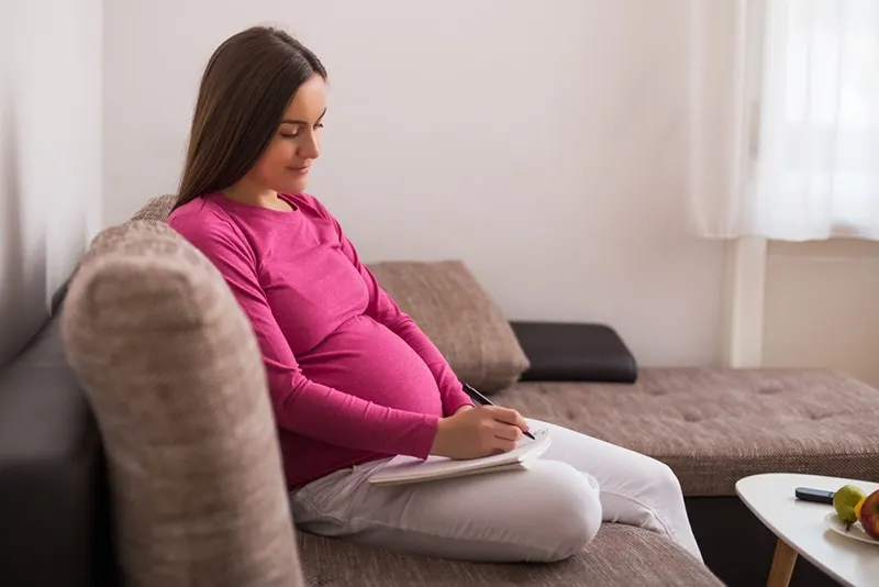 junge schwangere Frau, die im Wohnzimmer sitzt und einen Geburtsplan auf Papier schreibt