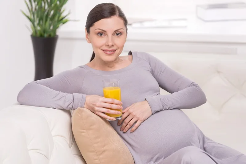 lächelnde schwangere Frau, die auf der Couch mit Saft in der Hand sitzt