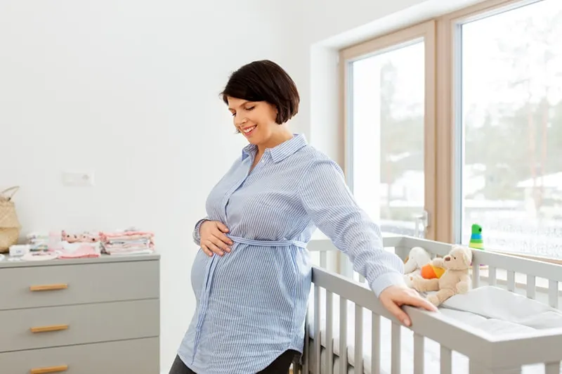 lächelnde schwangere Frau, die nahe dem Babybett im Schlafzimmer steht