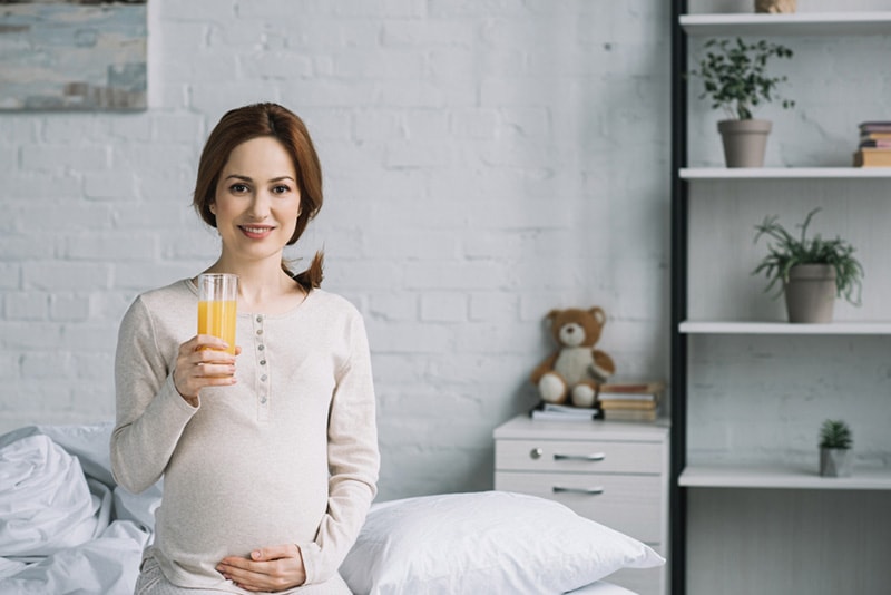 schwangere Frau, die im Schlafzimmer sitzt und Glas Saft hält