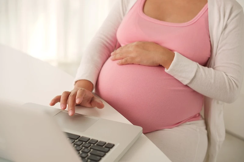 schwangere Frau, die im Stuhl mit Hand auf Bauch sitzt und mit anderen auf Laptop schreibt
