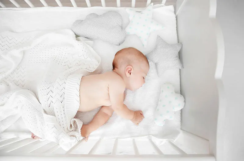 Baby schlafend im Kinderbett, zugedeckt mit Decke und mit Kissen herum