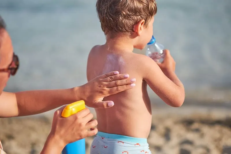 Eine junge Mutter sprüht Sonnencreme auf den Rücken eines Babys am Strand