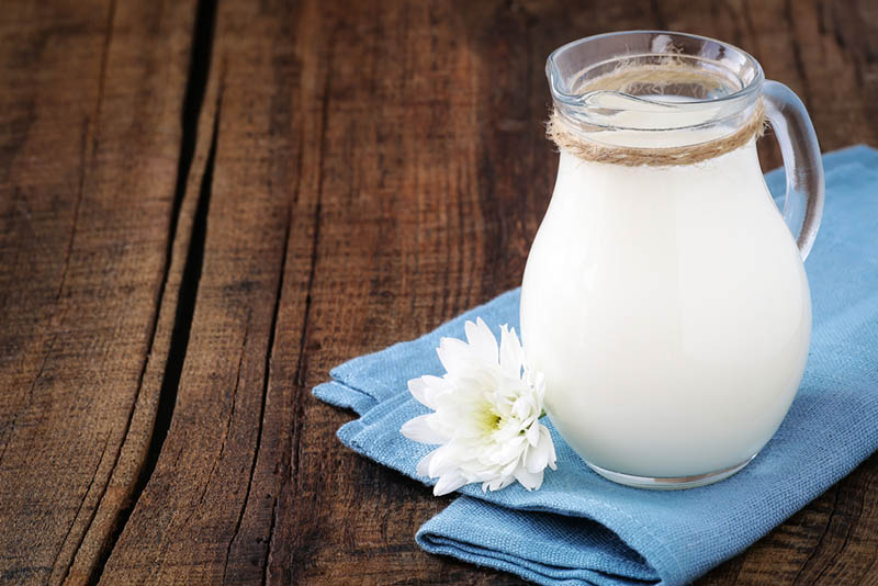 Frische Milch in einem Glaskännchen auf einer blauen Serviette mit einer Blume auf dem Holztisch