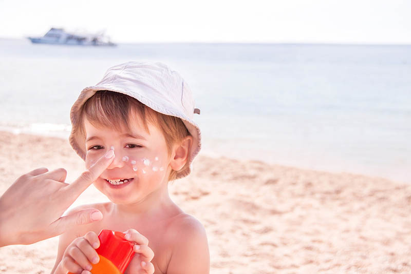 Mutter, die Sonnenschutzmittel auf lächelndes niedliches kleines Jungengesicht am Strand anwendet