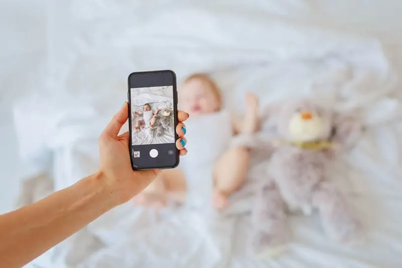 Mutter hält Smartphone und nimmt ein Bild von Baby auf dem Bett liegend mit Spielzeug