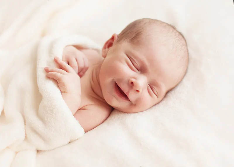  Neugeborenes Baby lachend, während es auf dem Bett mit weißer Decke bedeckt schläft