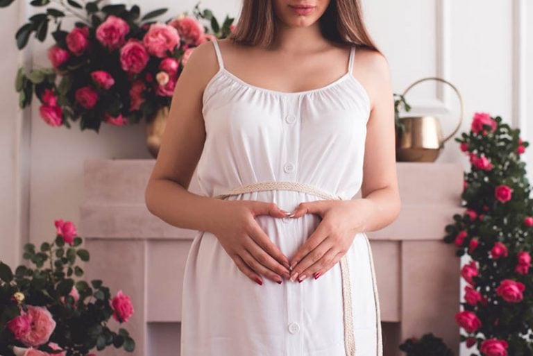 schöne schwangere Frau mit Blumen umgeben machen Herz mit Händen auf Bauch