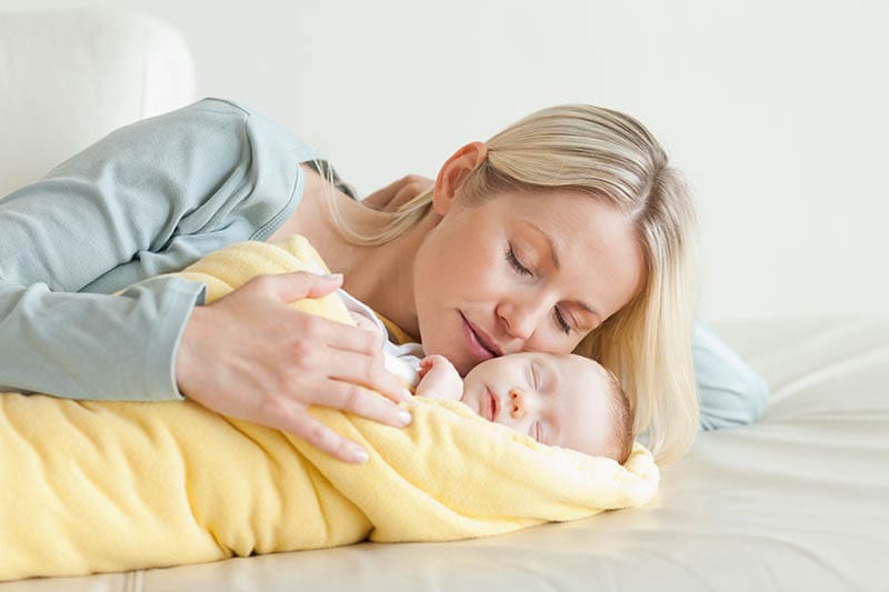 blonde Frau küsst sanft ihr schlafendes Baby auf dem Bett