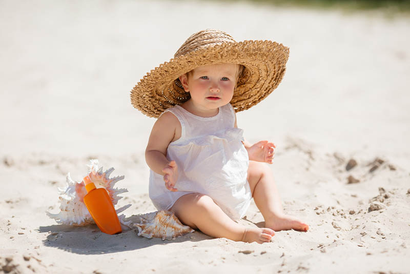 entzückendes kleines Mädchen mit Hut, der im Sand neben dem Sonnenschutz sitzt