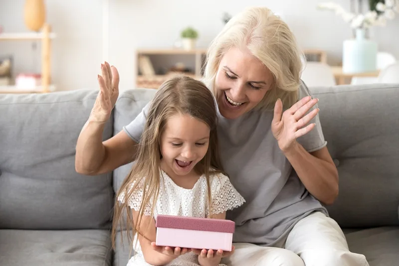 glückliche Großmutter überrascht ihre kleine Enkelin mit Geschenk im Wohnzimmer