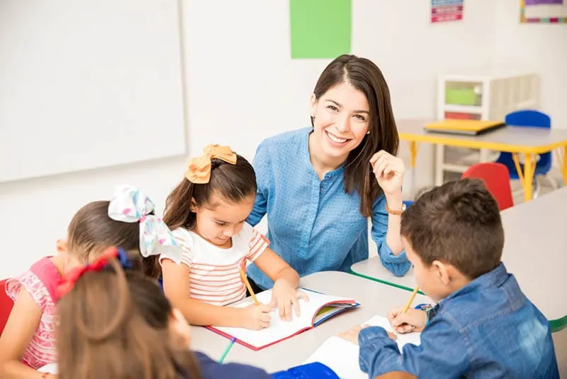 glückliche Lehrerin posiert, während Kinder in einem Notizbuch im Kindergarten schreiben
