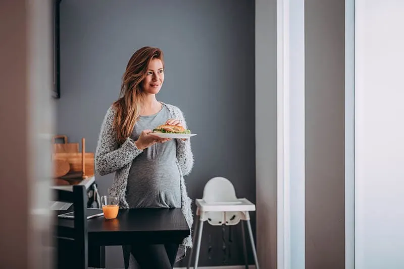 glückliche schwangere Frau isst ein Sandwich mit Schinken in der Küche am Fenster