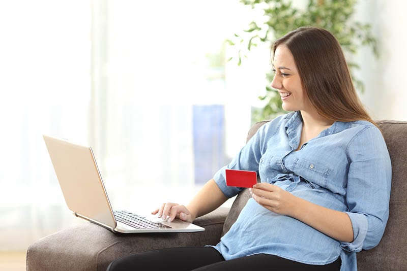glückliche schwangere Frau sitzt auf der Couch mit Laptop und eine Kreditkarte zu tun einige Online-Shopping