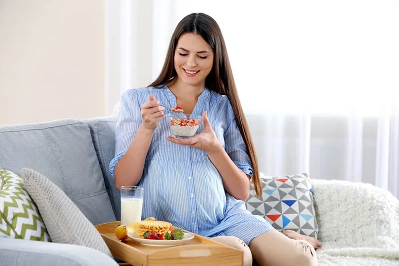 glückliche schwangere Frau sitzt auf der Couch zu Hause und hat ein Frühstück
