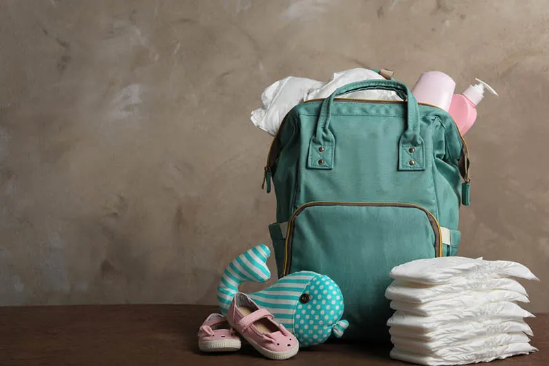 grüne Tasche mit Babywindeln und kleinen Schuhen auf dem Holztisch