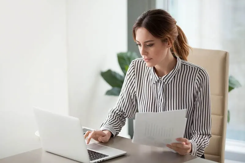 junge Frau sitzt am Tisch und tippt auf dem Laptop mit Papieren in der Hand
