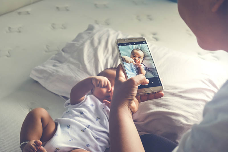 junge Mutter, die ihr Baby auf dem Bett mit Smartphone fotografiert