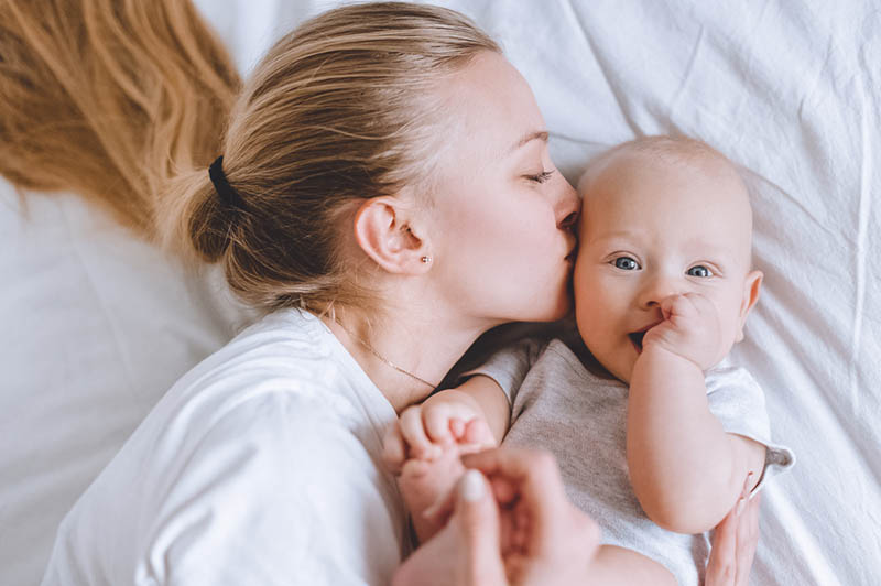 junge Mutter, die ihr süßes Baby auf dem Bett küsst und kuschelt