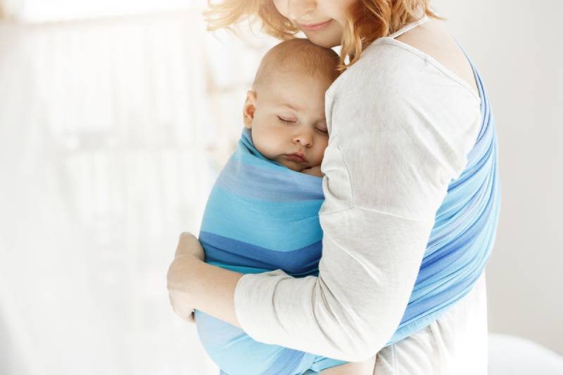 junge Mutter, die schlafendes Baby in einem blauen Träger trägt