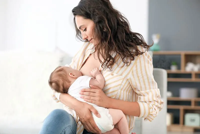 junge Mutter mit lockigem Haar stillt ihr Baby im Wohnzimmer