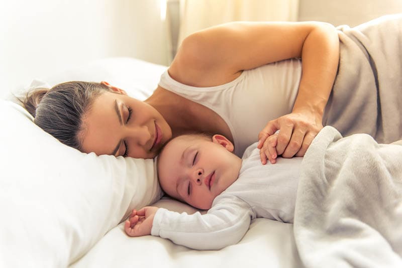 junge Mutter schlafend in Umarmung mit ihrem Baby auf dem Bett mit Blatt bedeckt