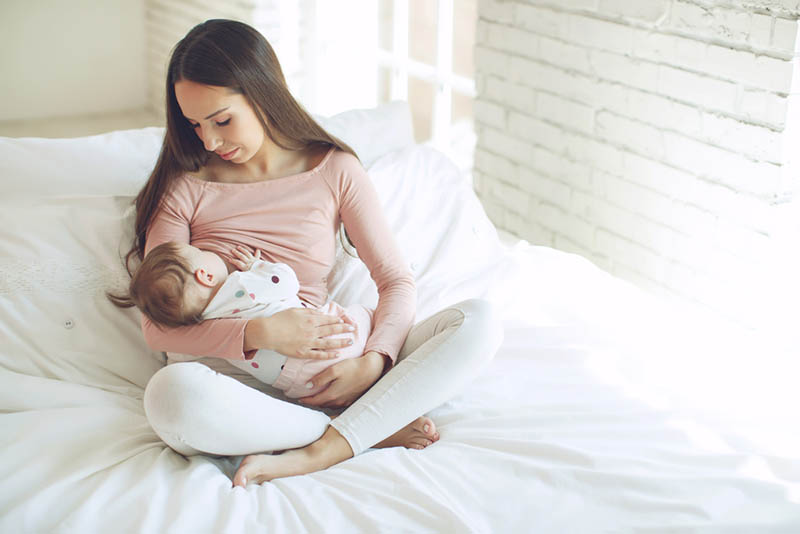 junge hübsche Mutter stillt Baby in Lotus Position auf dem Bett