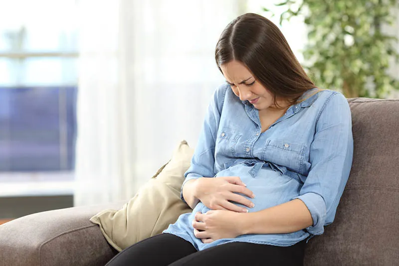 junge schwangere Frau sitzt auf der Couch und fühlt Schmerzen im Bauch