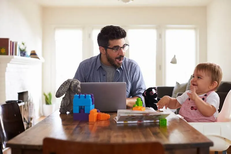 junger Vater arbeitet am Laptop, während seine kleine Tochter mit Spielzeug spielt