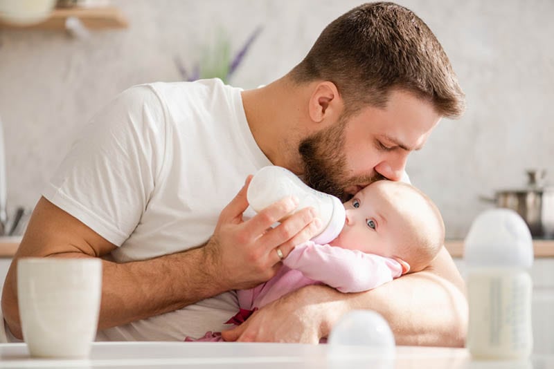 junger Vater füttert sein Baby mit der Flasche und küsst sie auf die Stirn