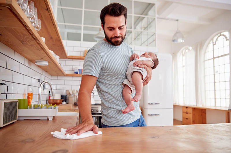 junger Vater hält schlafendes Baby und reinigt die Küchenoberfläche
