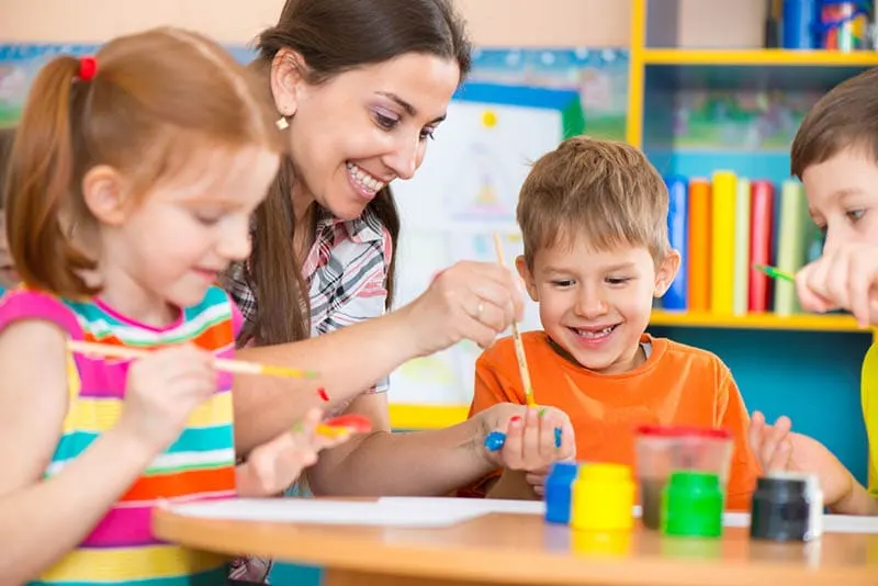 lächelnd Lehrer Malerei durch den Tisch mit Kindern im Kindergarten