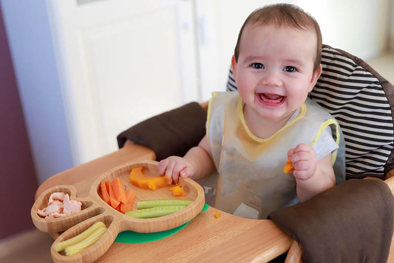 lächelndes Baby, das in einem Babystuhl sitzt und ein Gemüse isst
