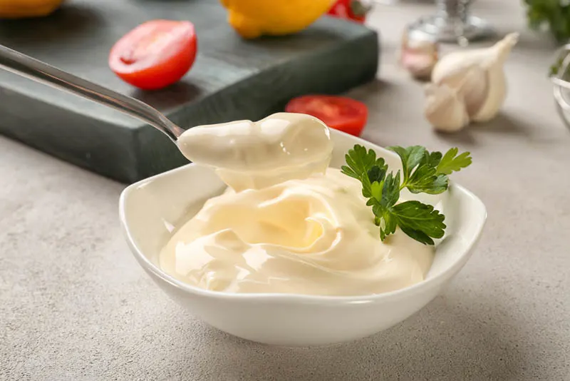 leckere Mayonnaise in weißer Schale und ein Löffel mit Zutaten auf dem Tisch