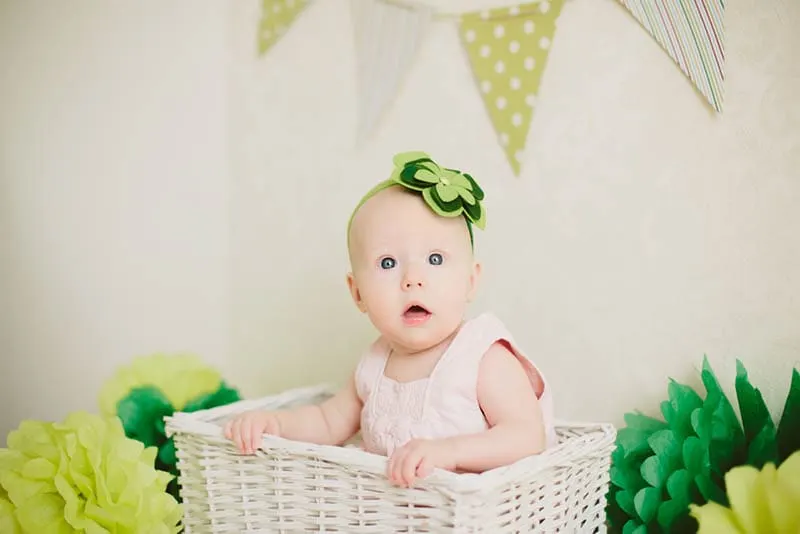 niedlichen Baby-Mädchen sitzt in einem hölzernen Korb mit grünem Papier Blumen
