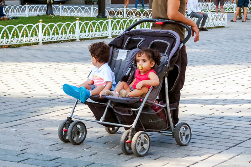 niedlichen Baby-Mädchen und ein Junge sitzen in einem Zwilling Kinderwagen mit Mutter im Park