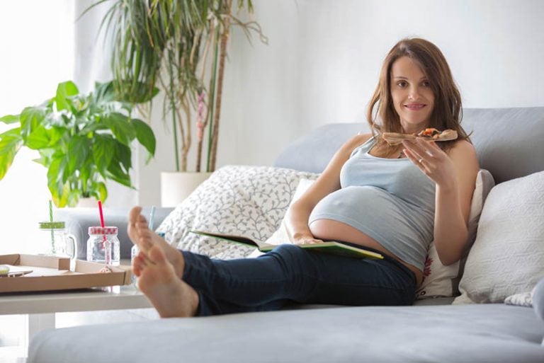 schwangere Frau liegt auf der Couch und isst eine Pizza